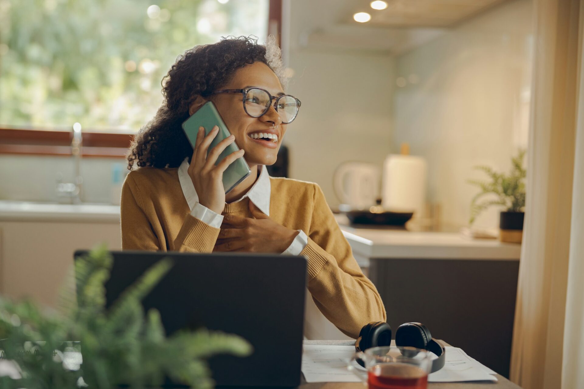 Female freelancer is talking phone while working on laptop from home sitting at kitchen